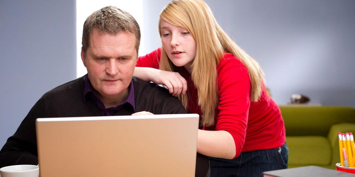 High school girl looking over her father's shoulder who is on a laptop
